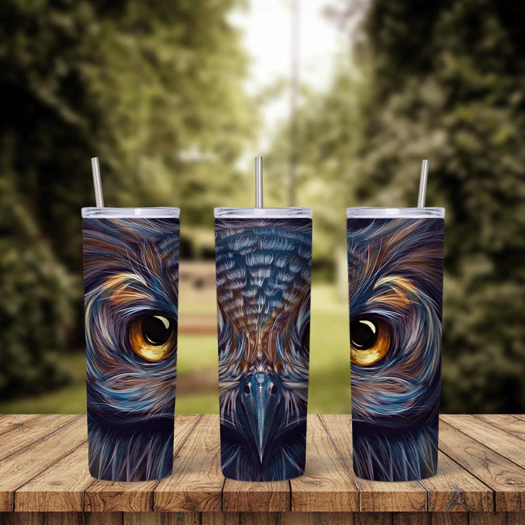 A 20 oz tumbler wrap with a painted close up of an owl face in blue, navy and orange feathers with glowing amber eyes. Sublimation design